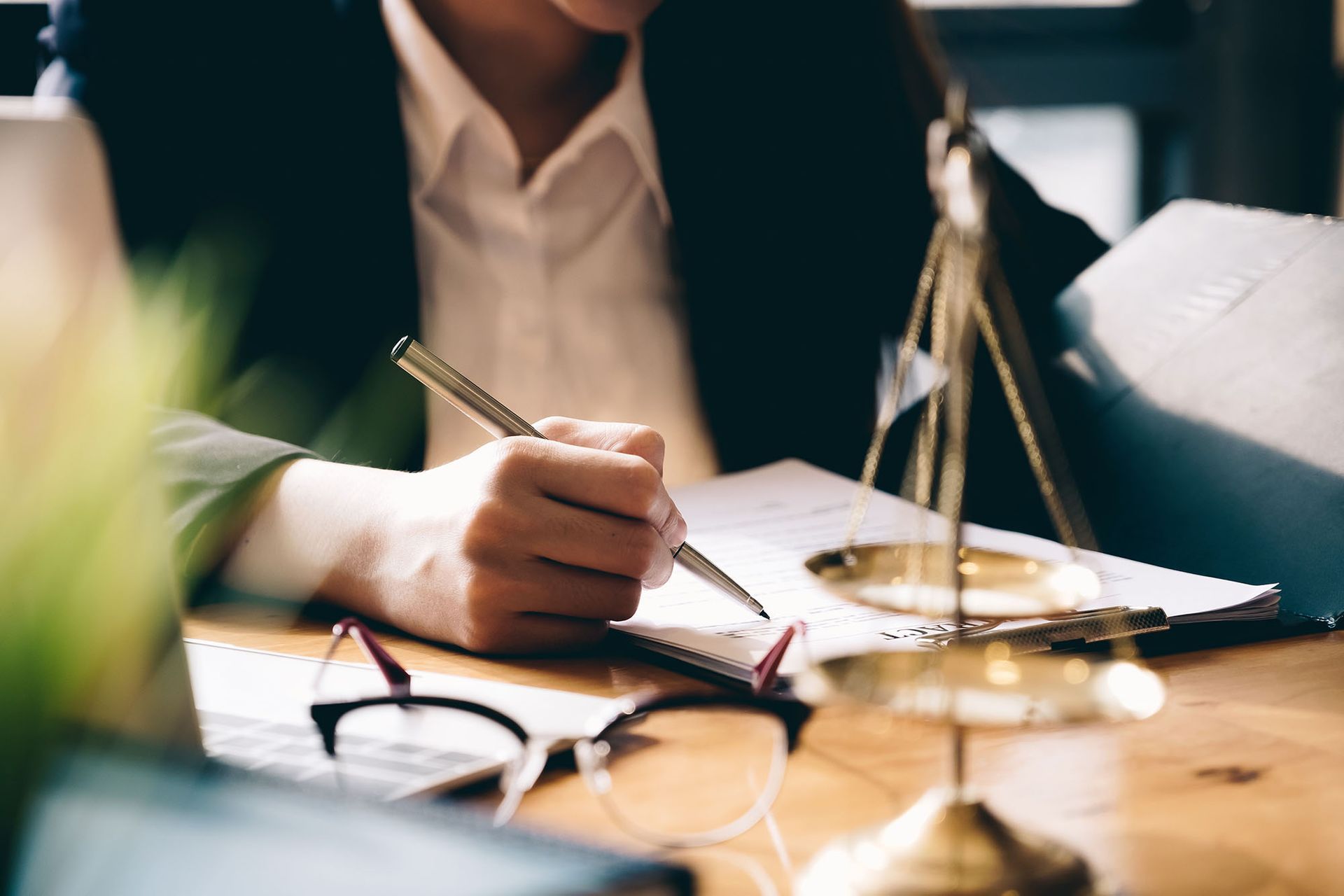 A person in a professional suit writes on paper at a desk featuring a brass scale of justice and eyeglasses.