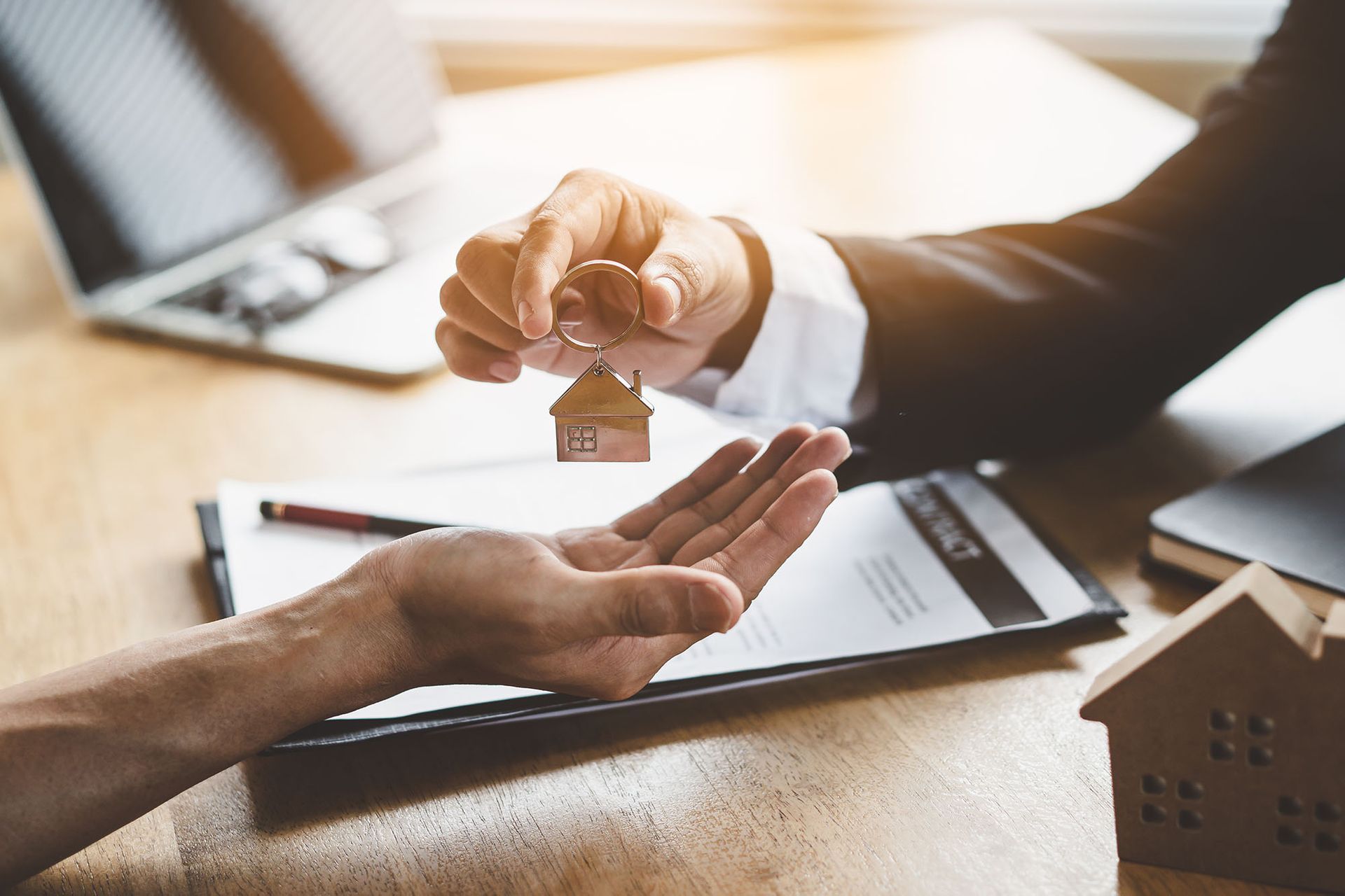 A person handing a house-shaped keychain to another person over a contract on a wooden desk.