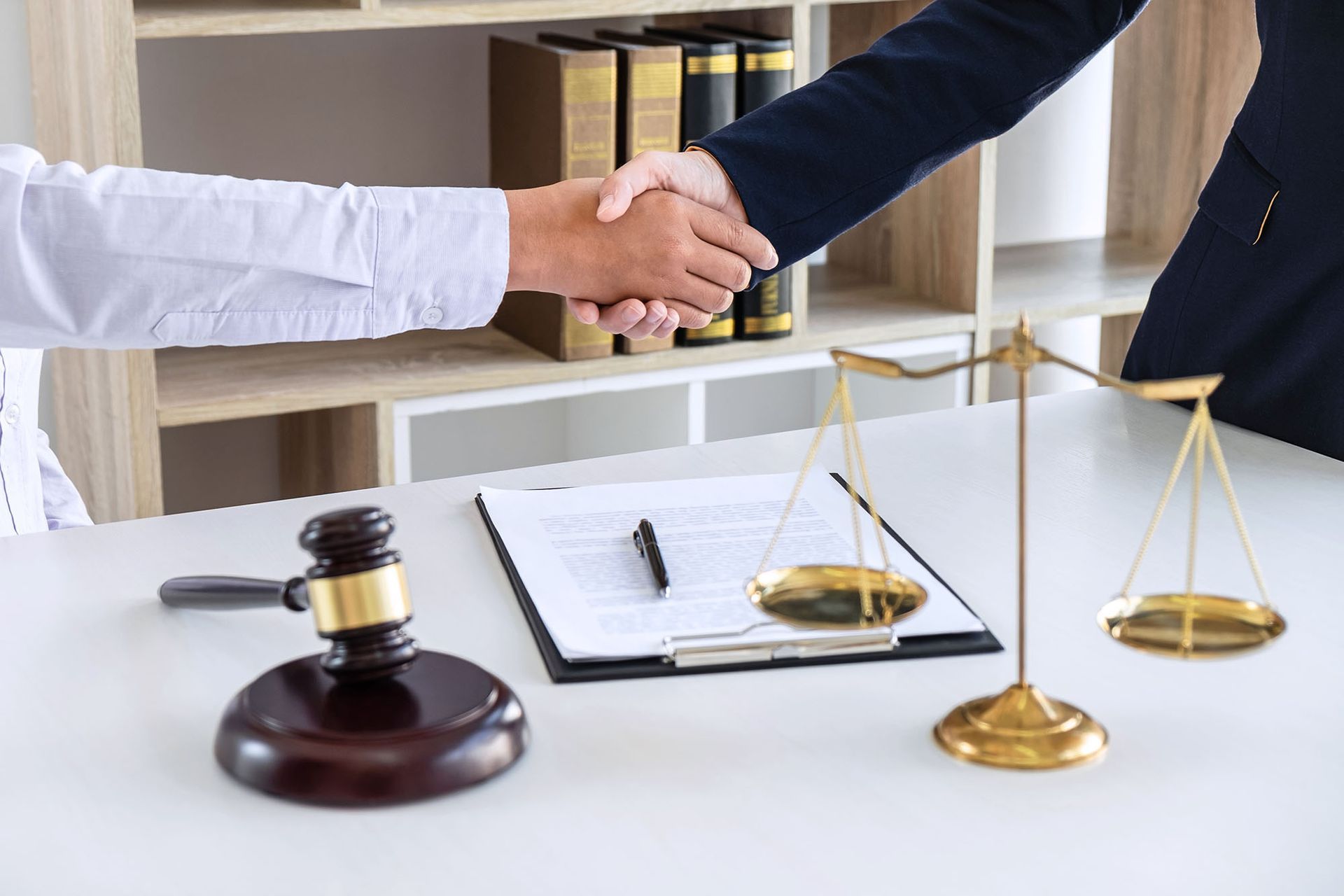 Two people in business attire shake hands over a desk with a gavel, legal document, and gold scales of justice.