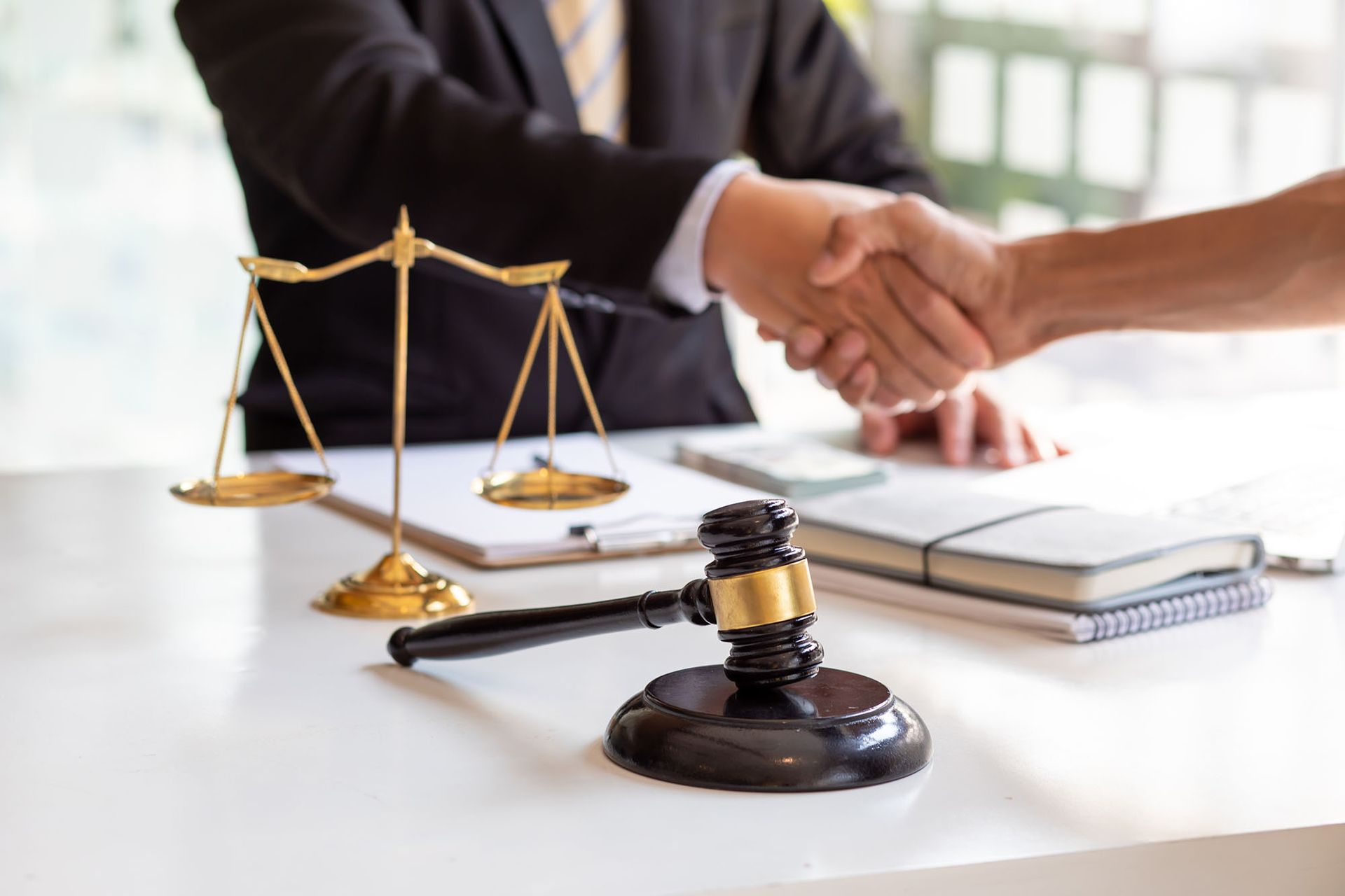 A person in a suit shakes hands with a client behind a legal scale and a wooden gavel on a desk.