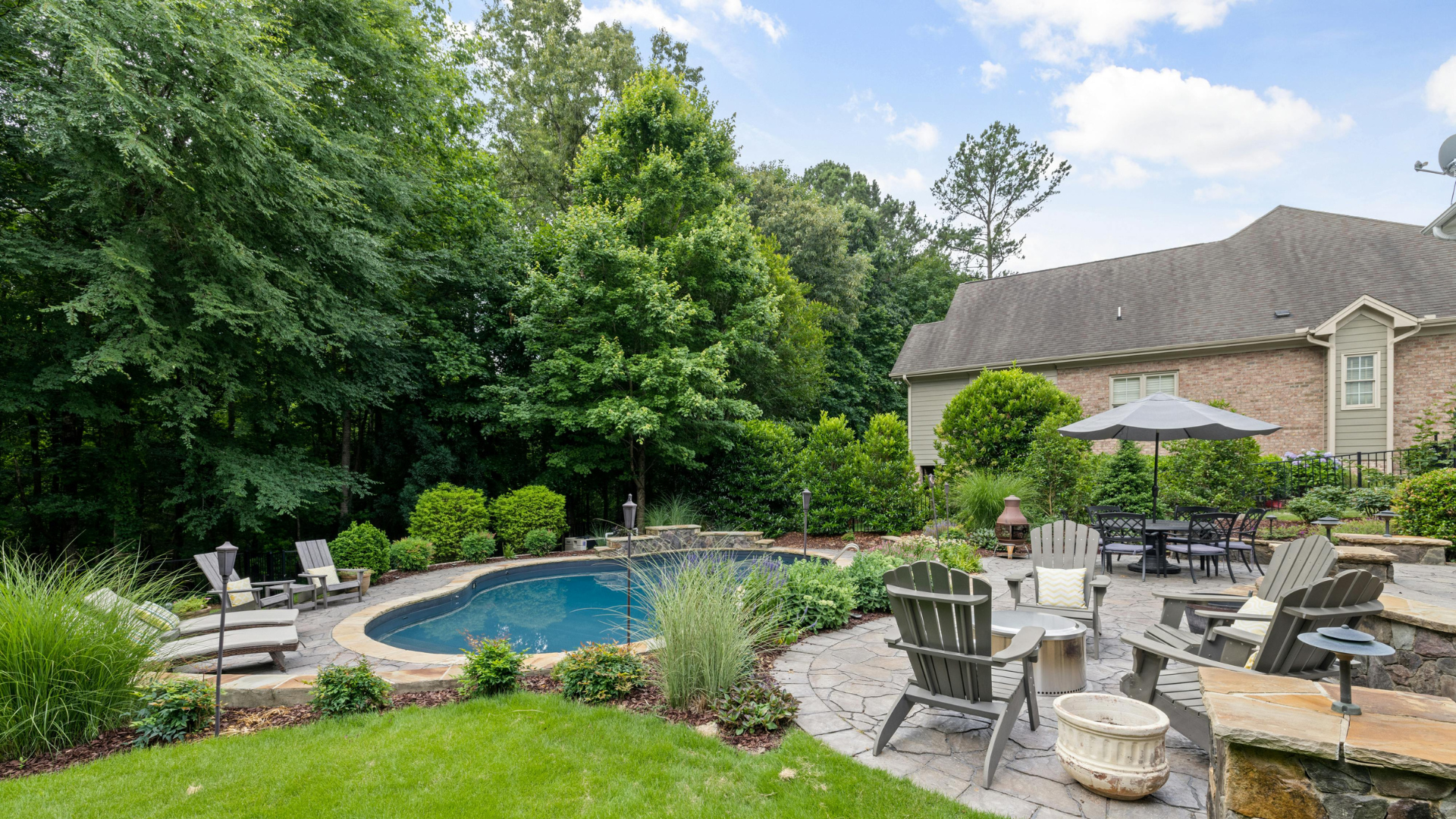 Backyard patio with a small pool, lounge chairs, and stone seating beside a house and trees