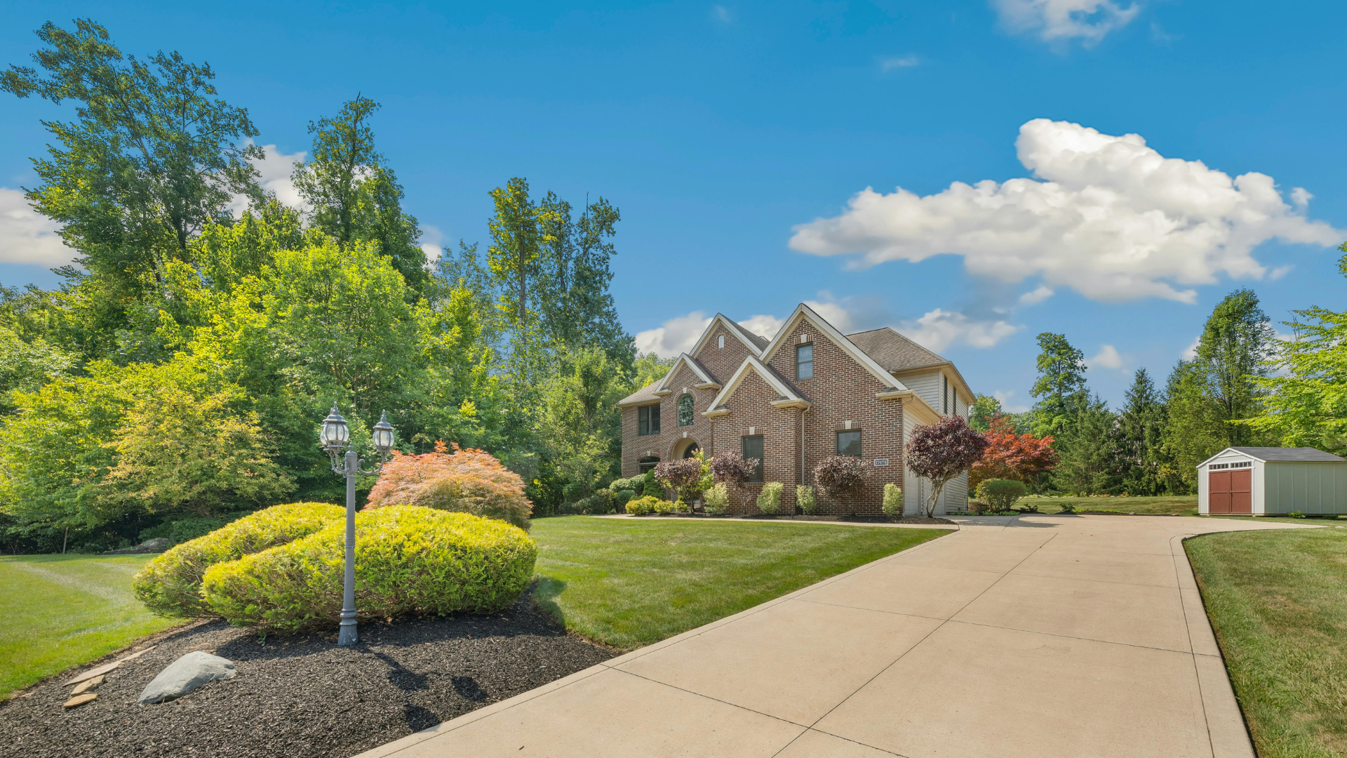 Suburban brick house with manicured lawn, driveway, and trees under a blue sky.