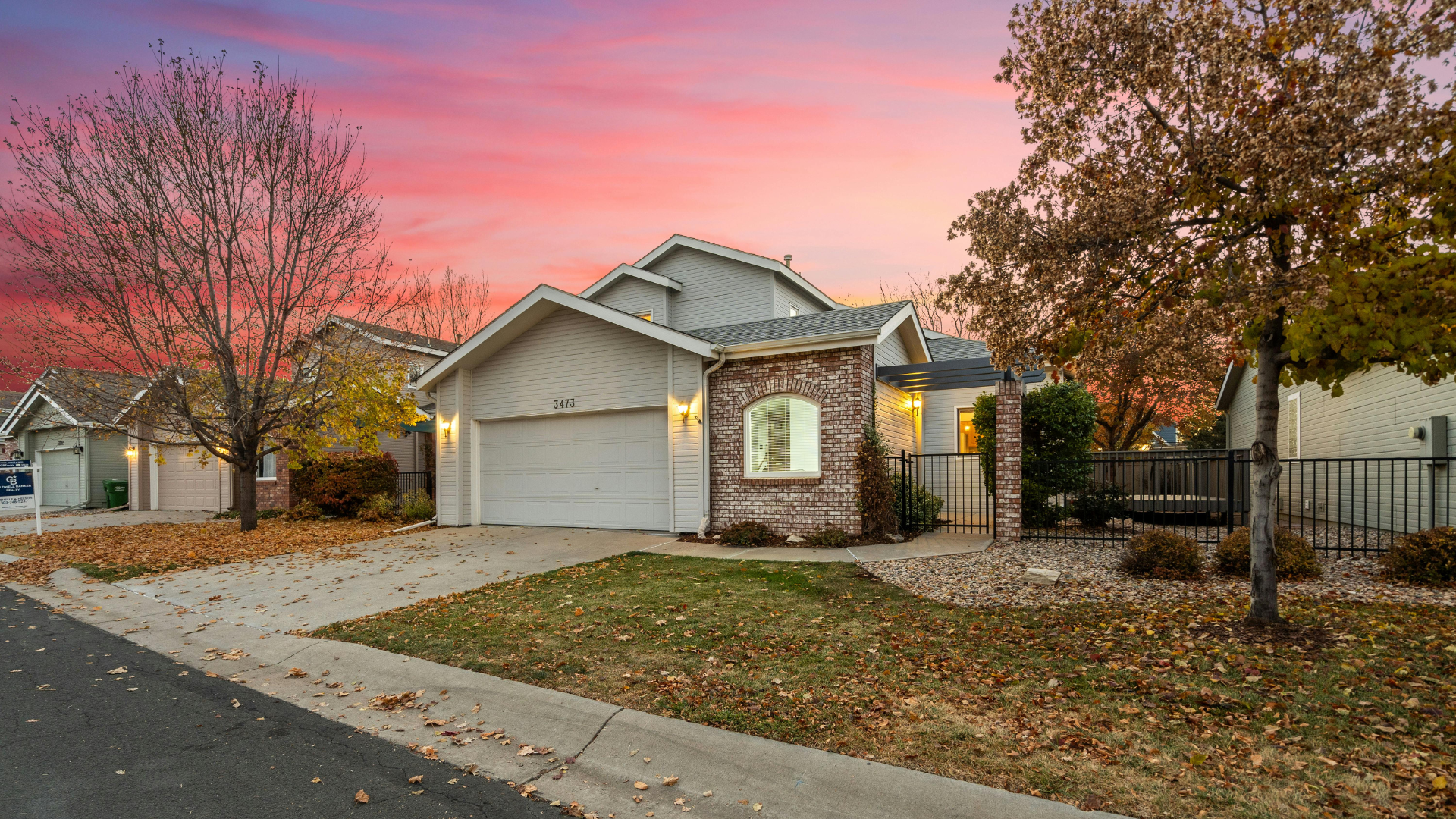 Suburban house with white garage and red brick facade at sunset, framed by trees and a leaf-covered yard.