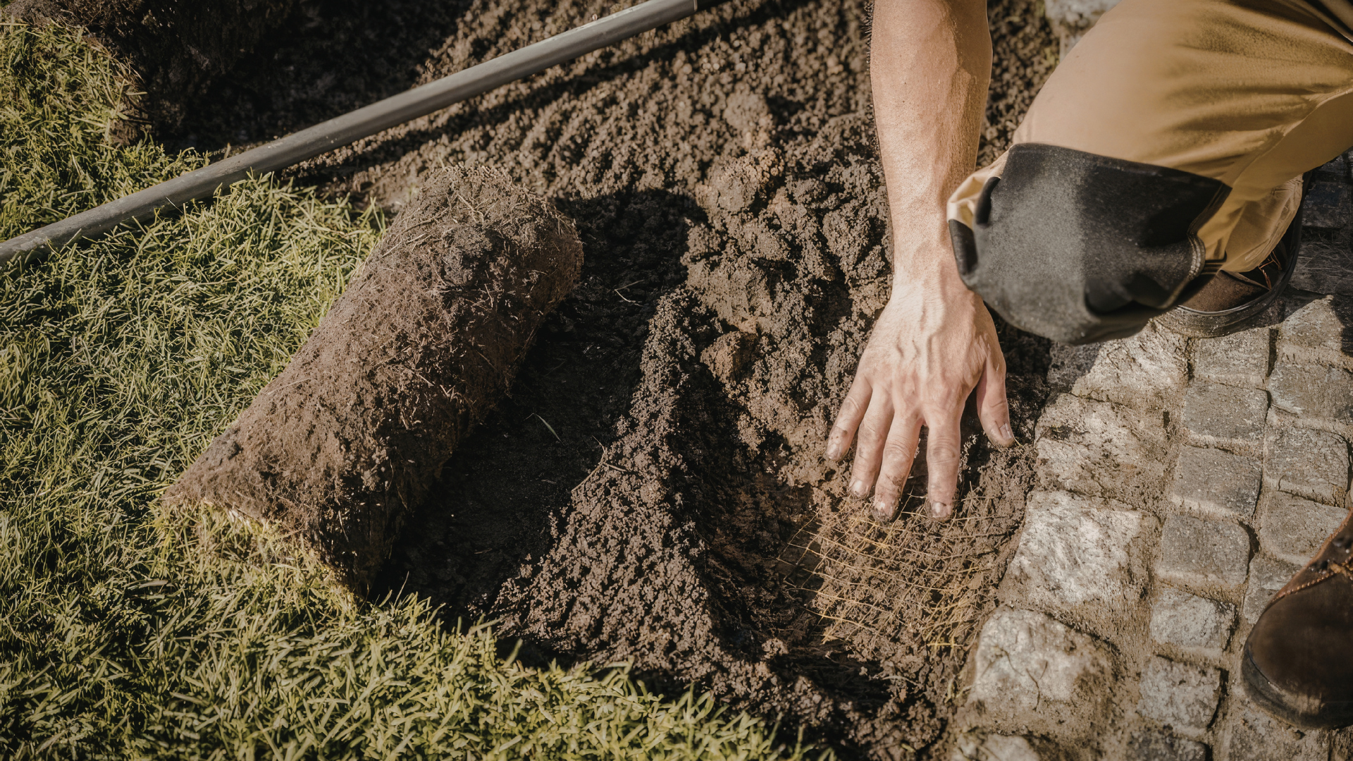 Person kneeling outdoors, digging soil beside a stone path with a shovel.