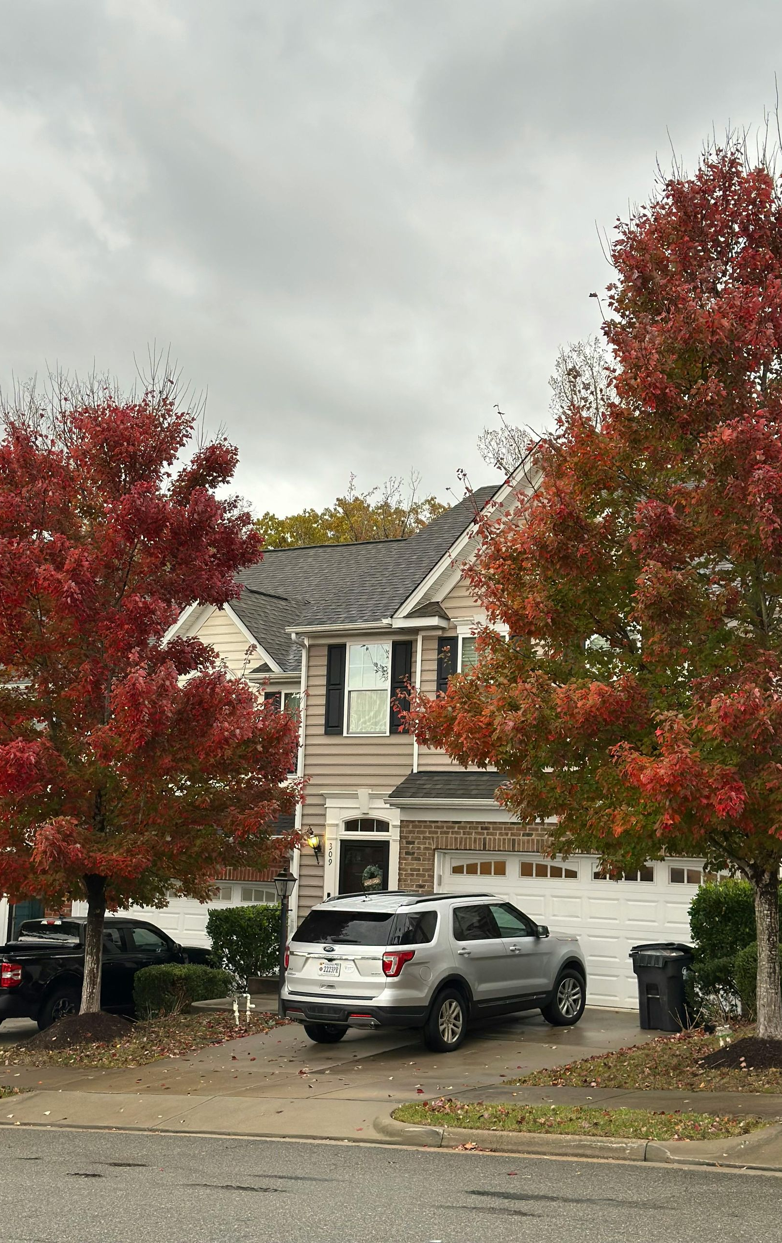 Two-story house with red-leafed trees in front; gray SUV parked in driveway under cloudy sky.