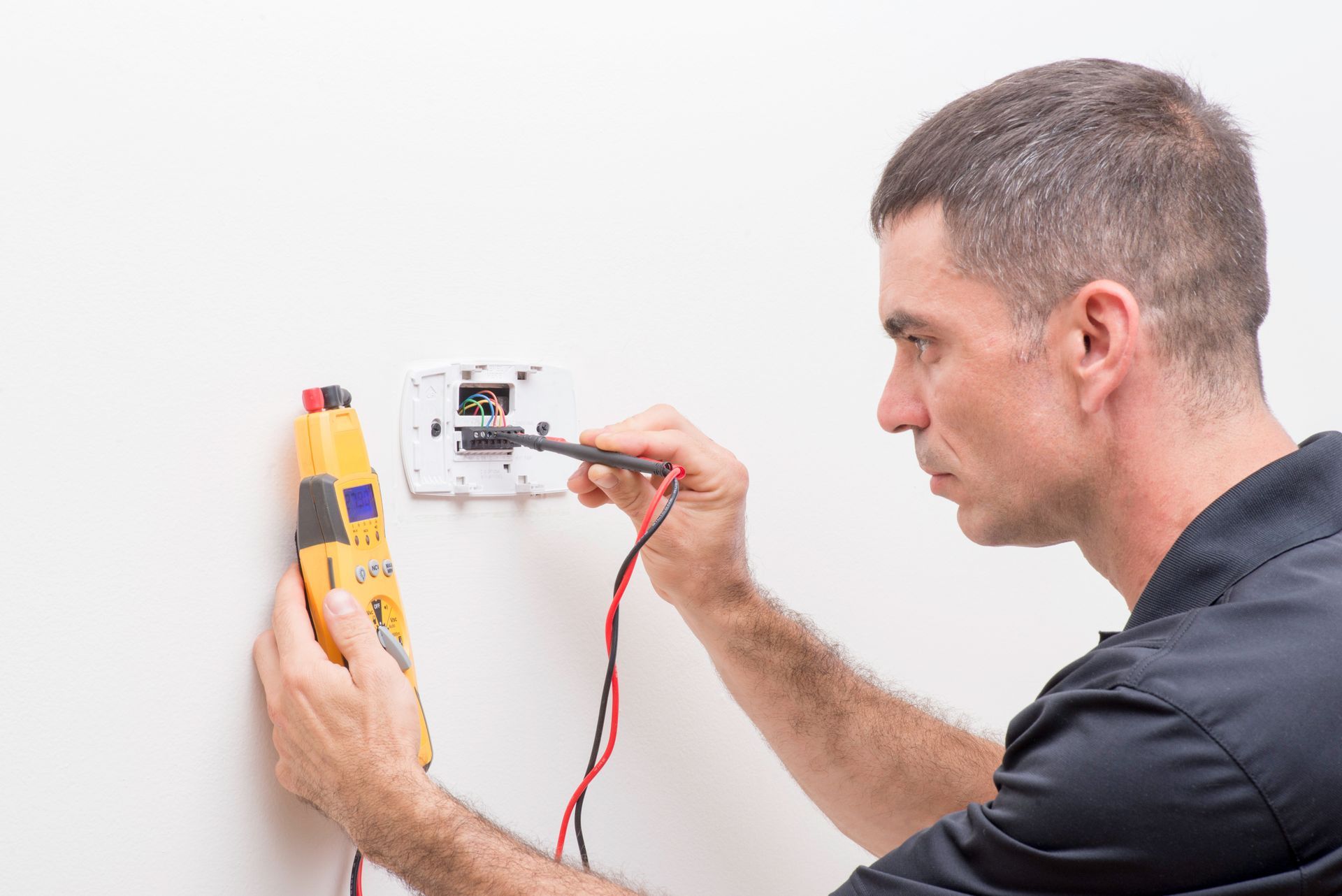Electrician testing thermostat wiring with multimeter on wall-mounted HVAC control panel inspection.