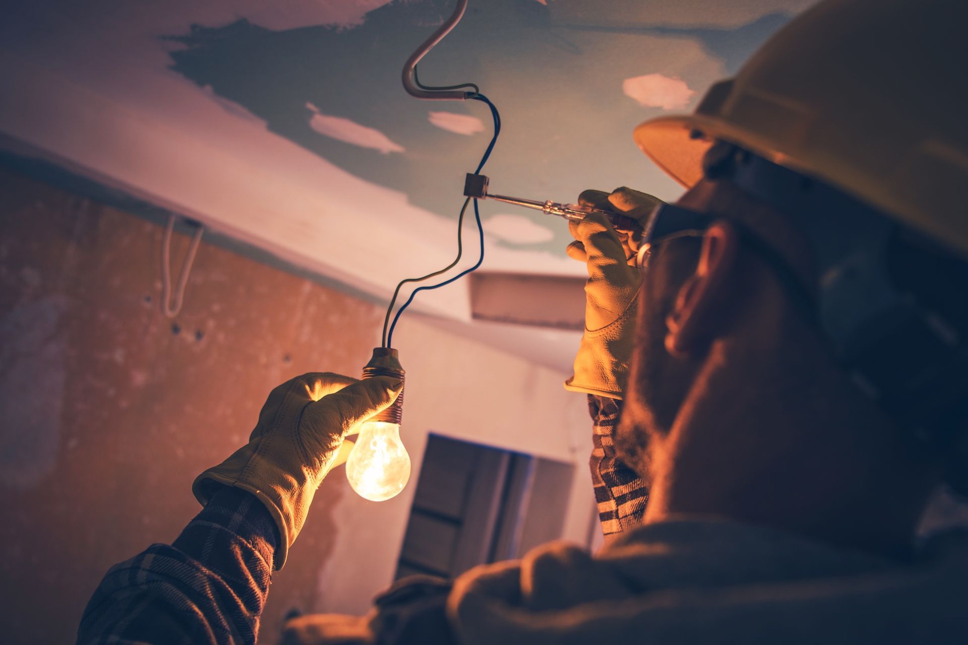Electrician wiring a ceiling light fixture using tools and safety gear.