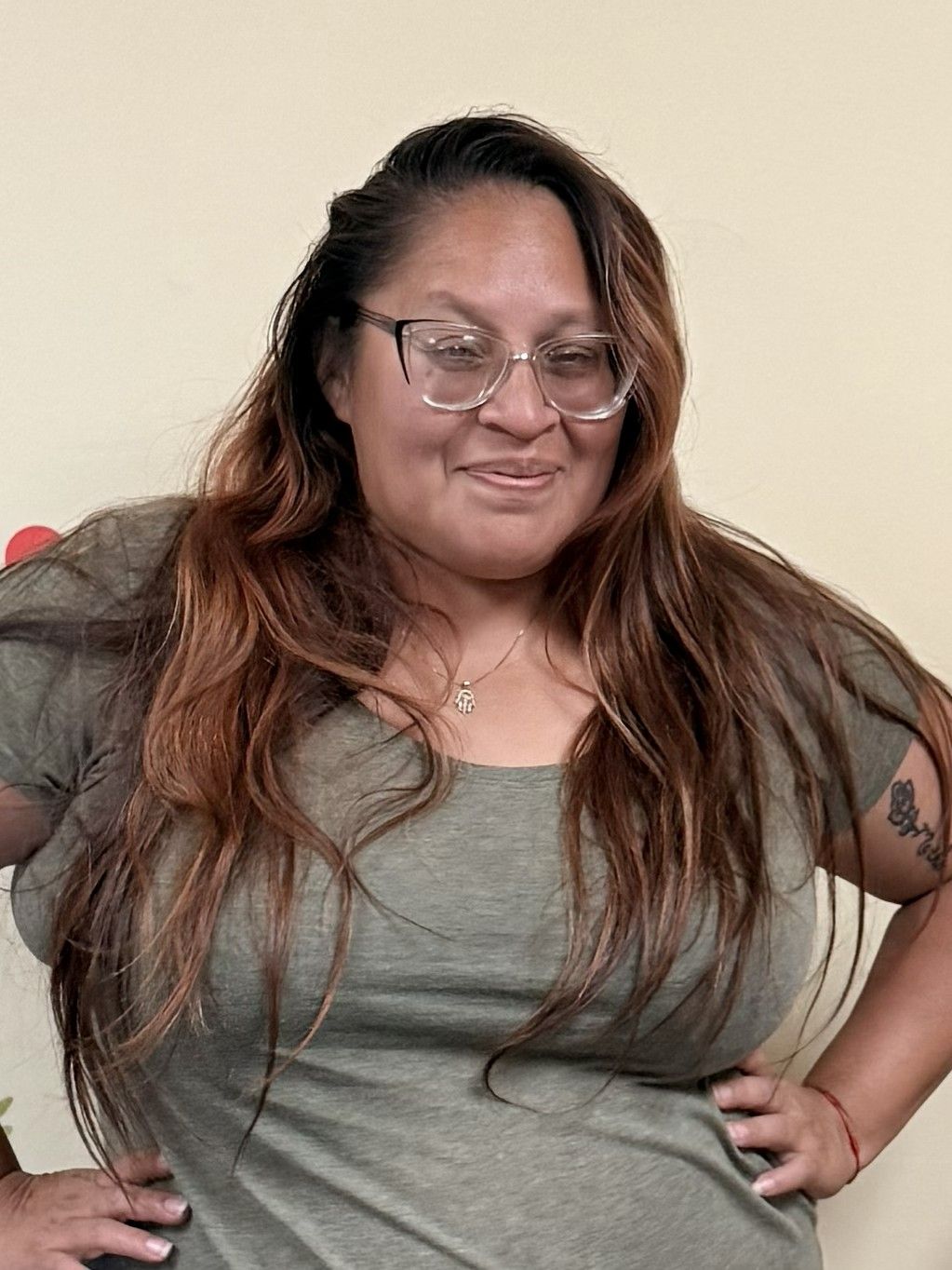 Smiling person in a gray shirt and maroon cardigan against a beige wall, with sunglasses hanging from the collar