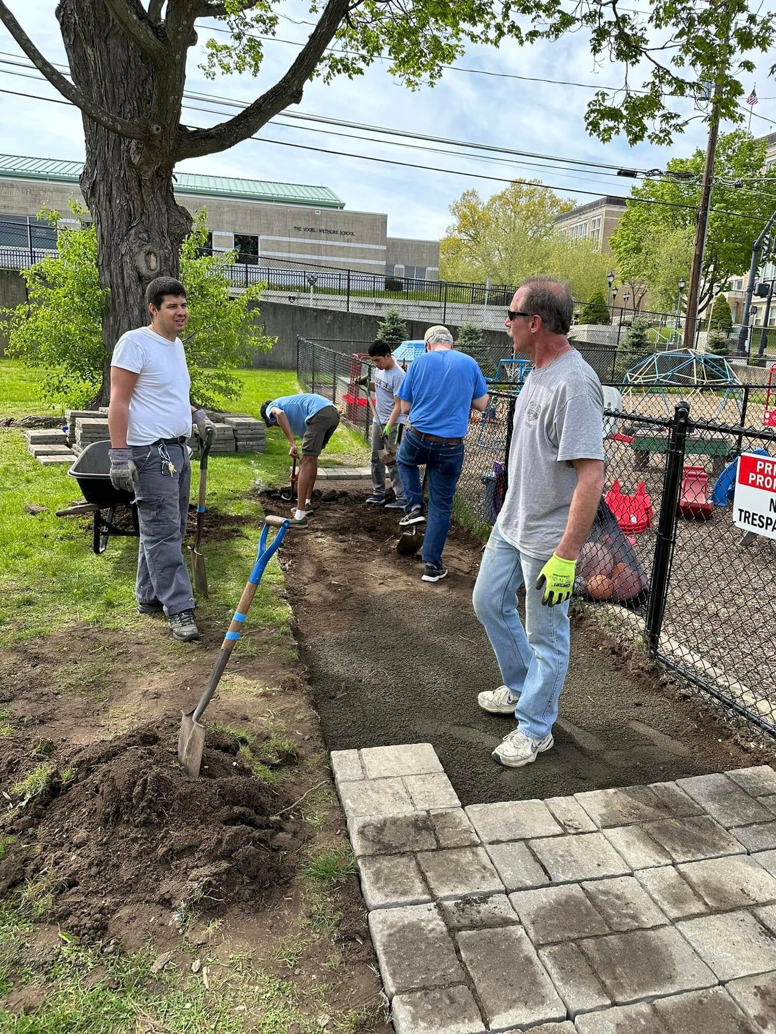 People digging a garden path beside a fence and tree in a park-like area.
