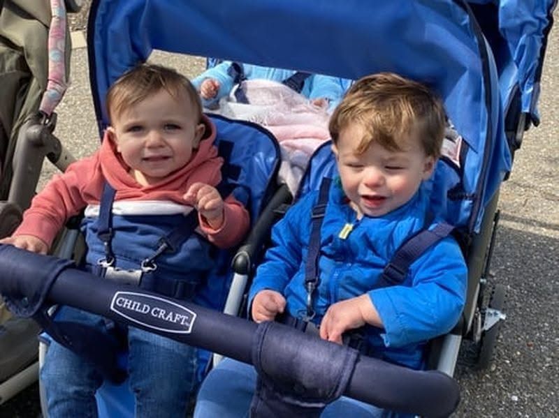 Two toddlers in a blue stroller, smiling outdoors with a black safety bar in front
