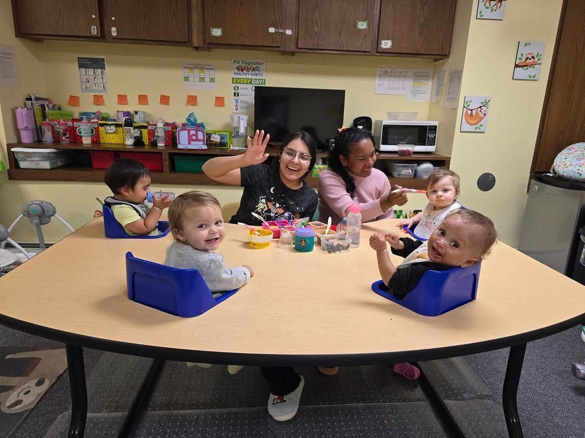 Children and adults at a daycare table, one adult waving, with snacks and toys in a classroom