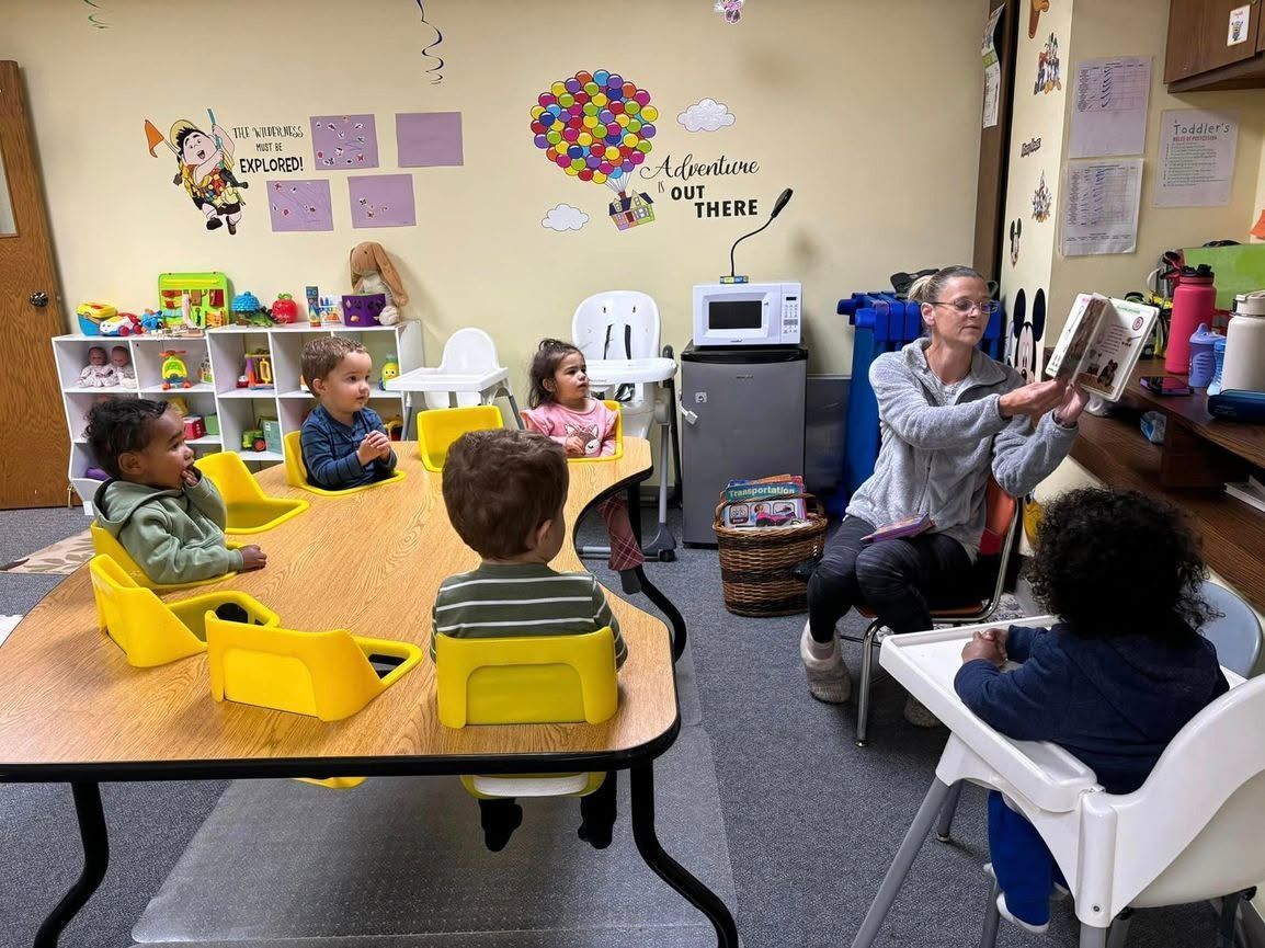 Children sit at a classroom table while a teacher works with one child near a printer.