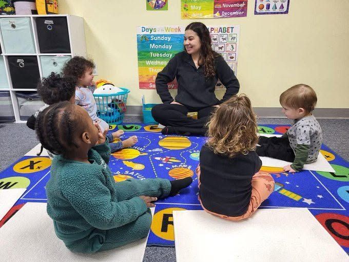 Teacher seated on a colorful classroom rug, leading a small circle time with children.