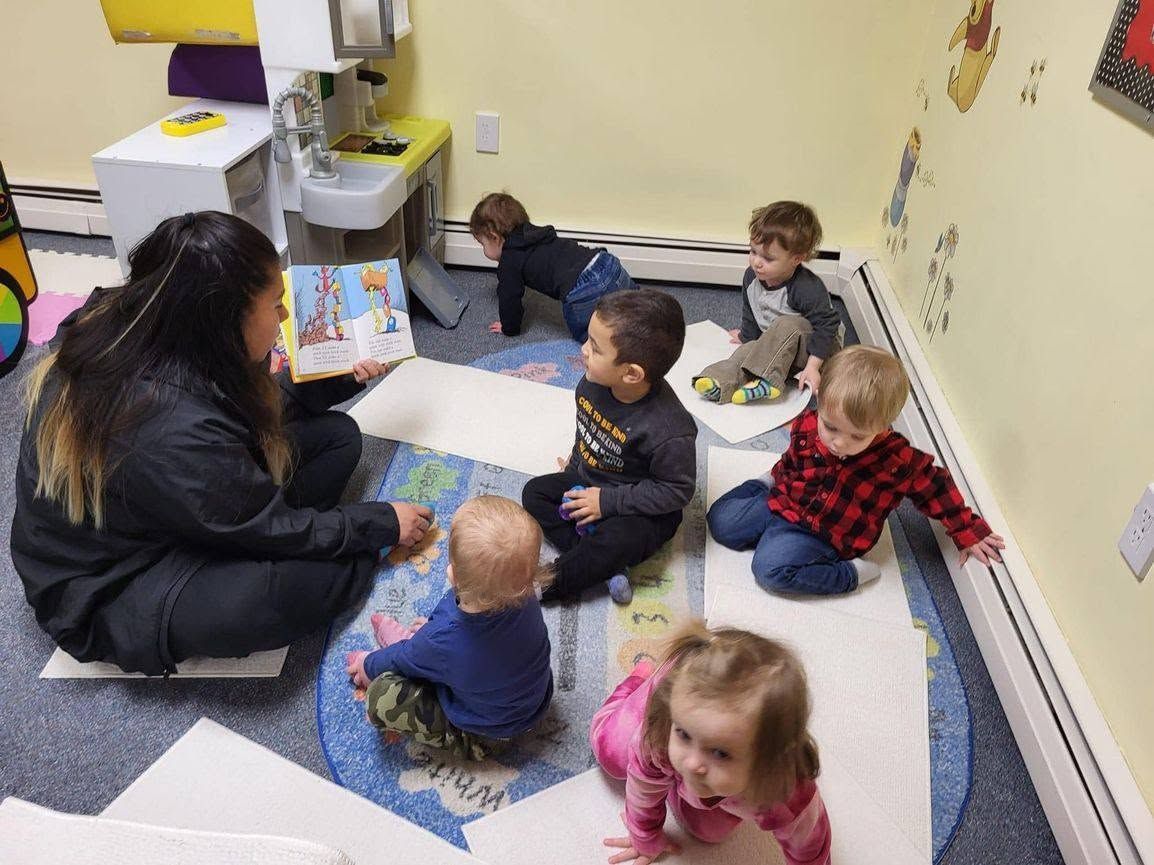 Caregiver reading to several children sitting on a classroom rug with books and toys.