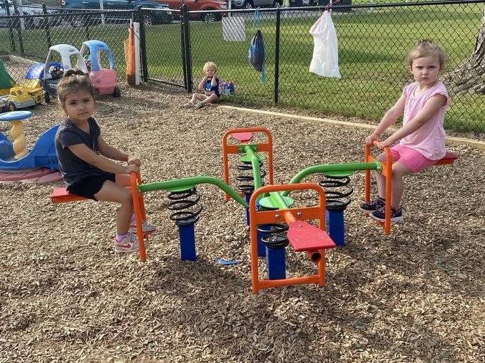 Two children playing on colorful spring riders in a playground with wood chips and a fence backdrop