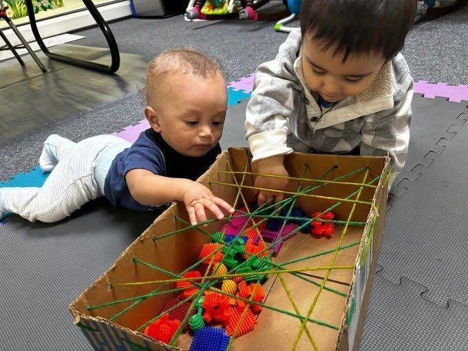 Two toddlers play with colorful pom-poms in a cardboard box maze on a padded floor.