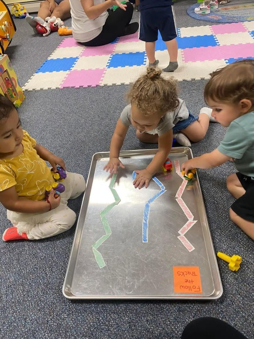 Children drawing on a lighted tray in a classroom play area with colorful foam mats.