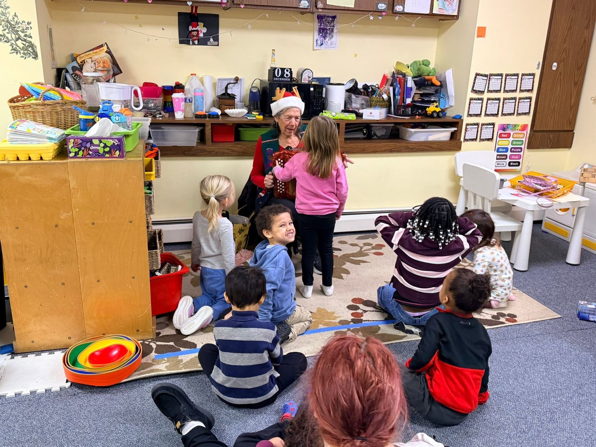 Children sitting on a classroom rug while a teacher stands by the sink and counter area