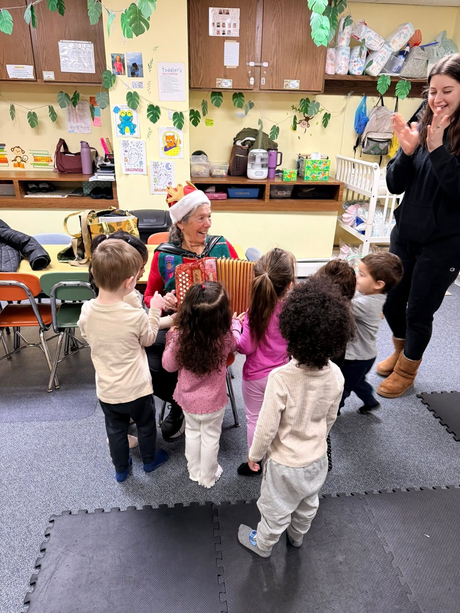 Children gathered around a teacher in a colorful classroom, listening and raising hands.