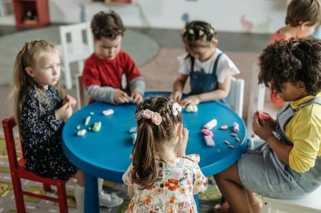 Children seated around a blue table, playing with small colorful objects in a classroom