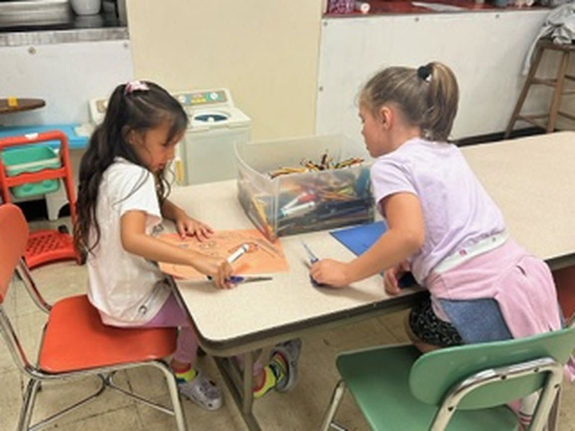 Two children sit at a table drawing with crayons in a classroom.