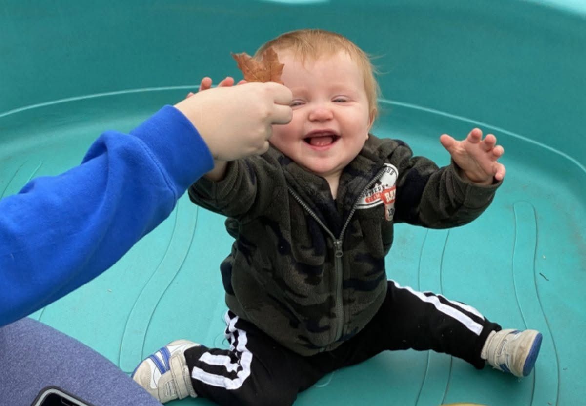 Smiling baby in a black jacket sitting on a teal surface, reaching toward a hand in a blue sleeve
