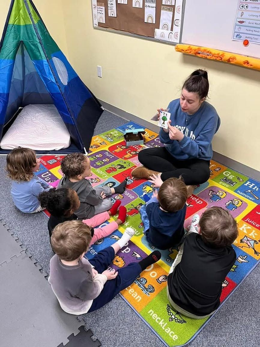 Teacher reading to children seated on a colorful classroom rug, with a play tent nearby.