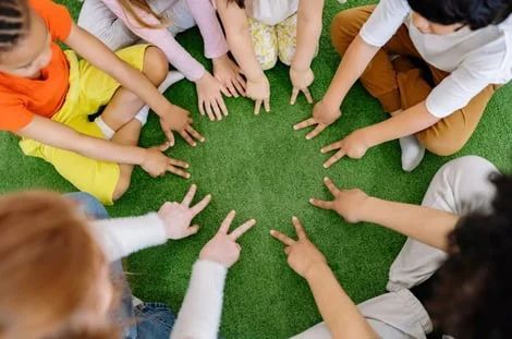 Children sitting in a circle on grass, pointing inward with hands together.