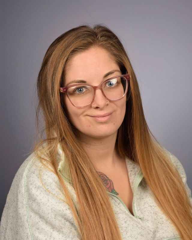 Smiling person in a gray shirt and maroon cardigan against a beige wall, with sunglasses hanging from the collar