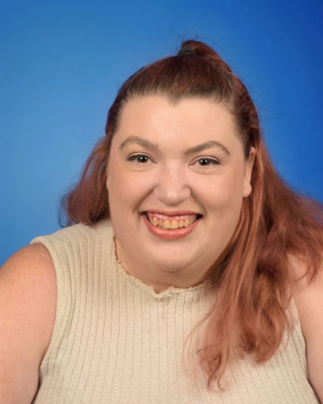 Smiling person in a gray shirt and maroon cardigan against a beige wall, with sunglasses hanging from the collar