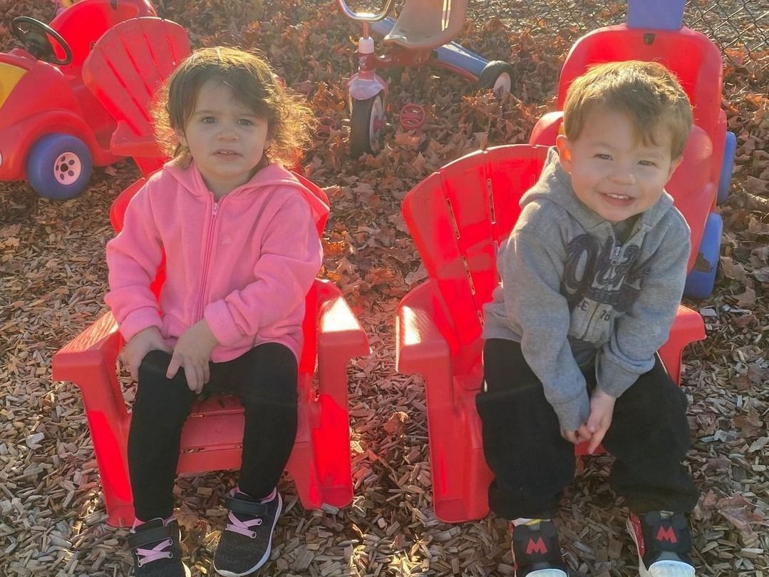 Two children sitting on red chairs outdoors among fallen leaves, one in a pink jacket and one in a gray jacket, smiling