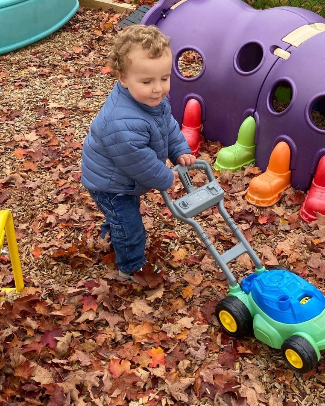 Child pushing a green toy lawn mower on a leaf-covered playground near a purple play structure