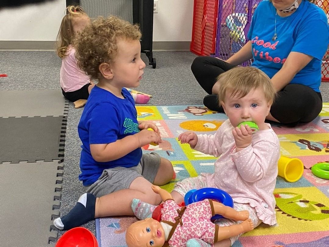 Two toddlers play with toys on a colorful mat while an adult sits behind them.