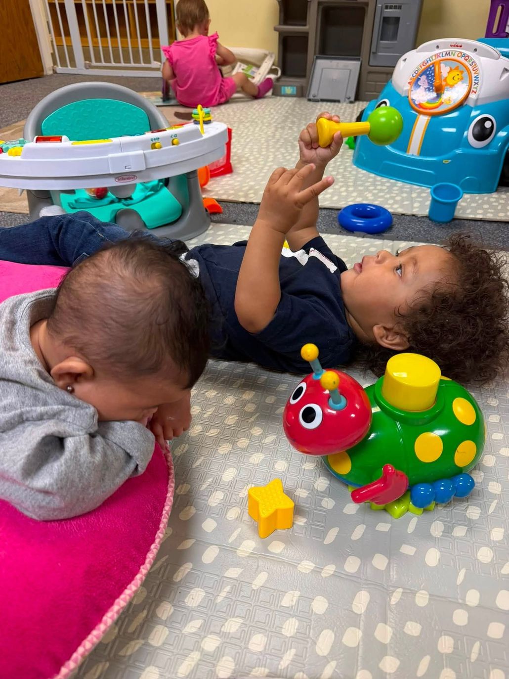 Smiling baby in a black jacket sitting on a teal mat while an adult hand touches their nose