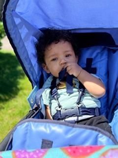 Child sitting in a blue stroller, holding a hand near their mouth, under a blue canopy.