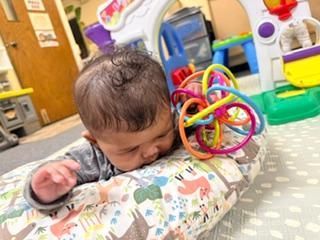 Sleeping baby resting on a patterned cushion beside a colorful ring toy in a playroom