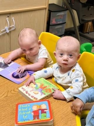 Two babies sitting at a daycare table with colorful books and toys, one reaching for a toy panel