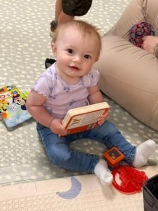 Baby sitting on a patterned play mat, holding a book and smiling beside toys.