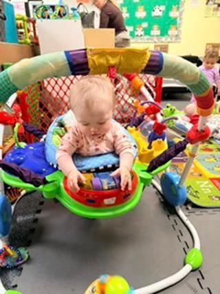 Baby sitting in a colorful activity bouncer indoors, looking down with toys around them