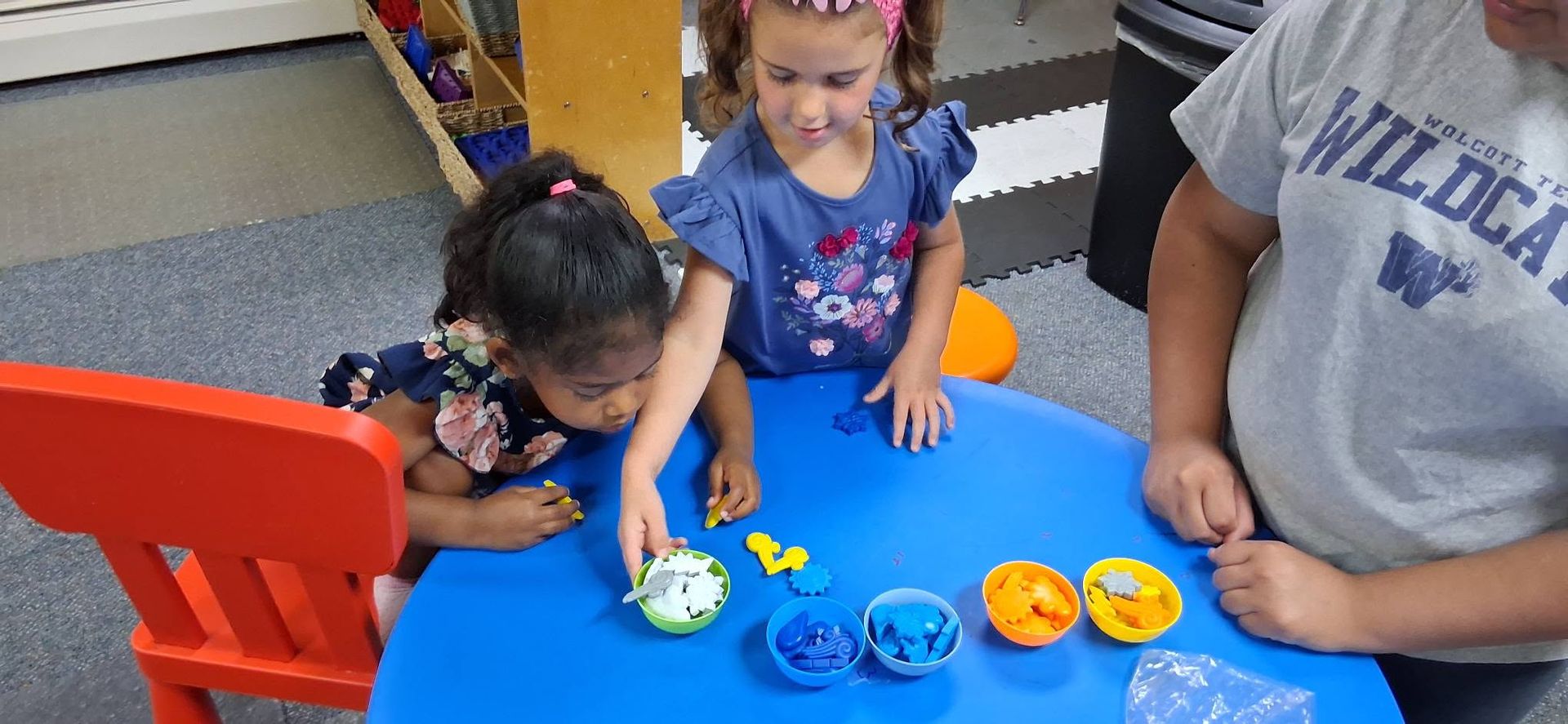 Children playing with colorful toy bowls and cups at a blue table in a classroom.
