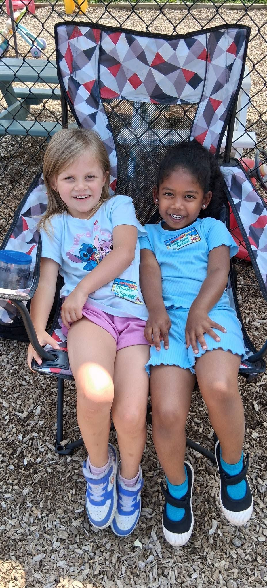 Two children sitting in folding chairs outdoors, smiling, with a quilt hanging behind them.