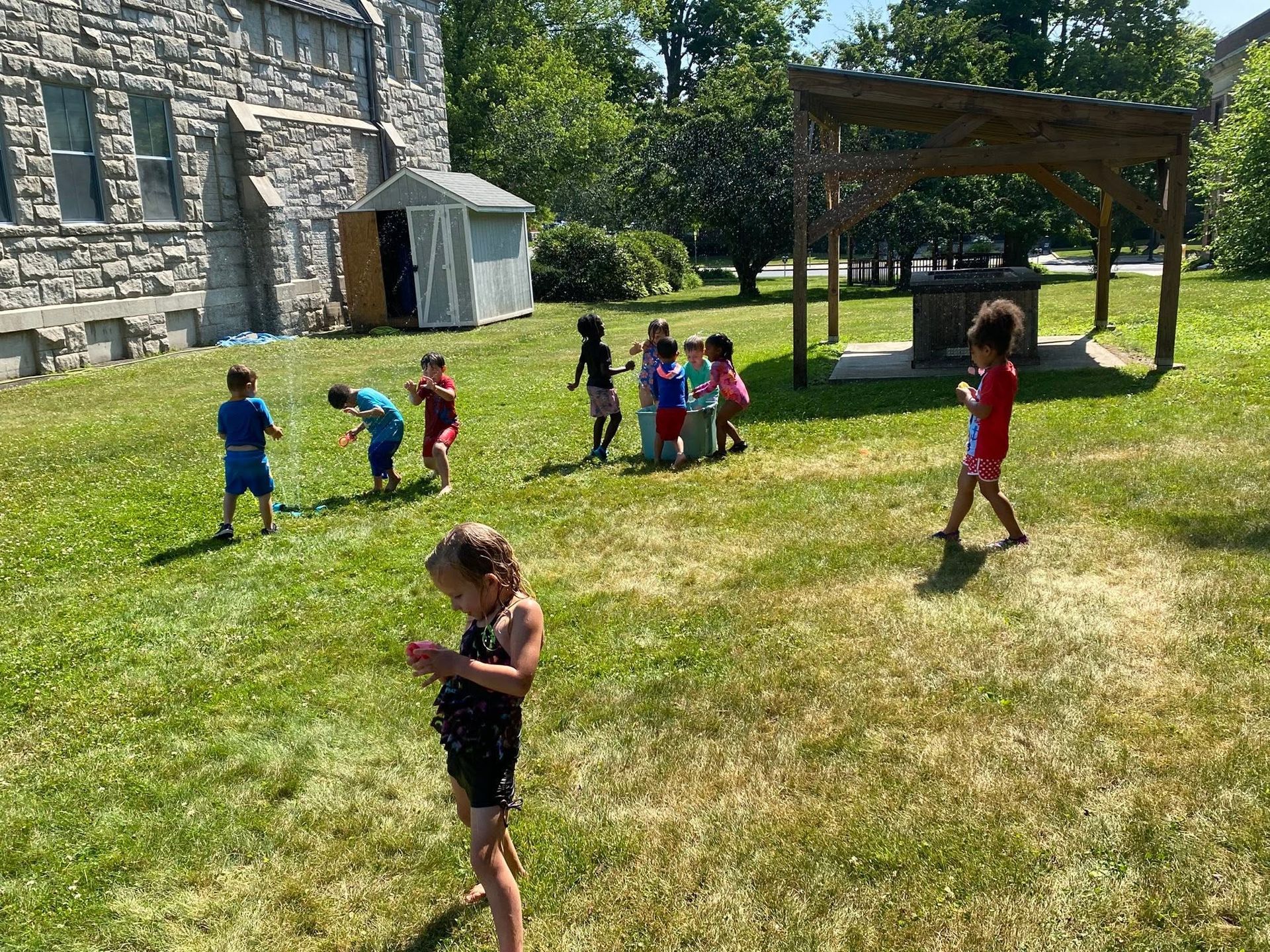 Children playing on a sunny lawn beside a stone building and a wooden pavilion.