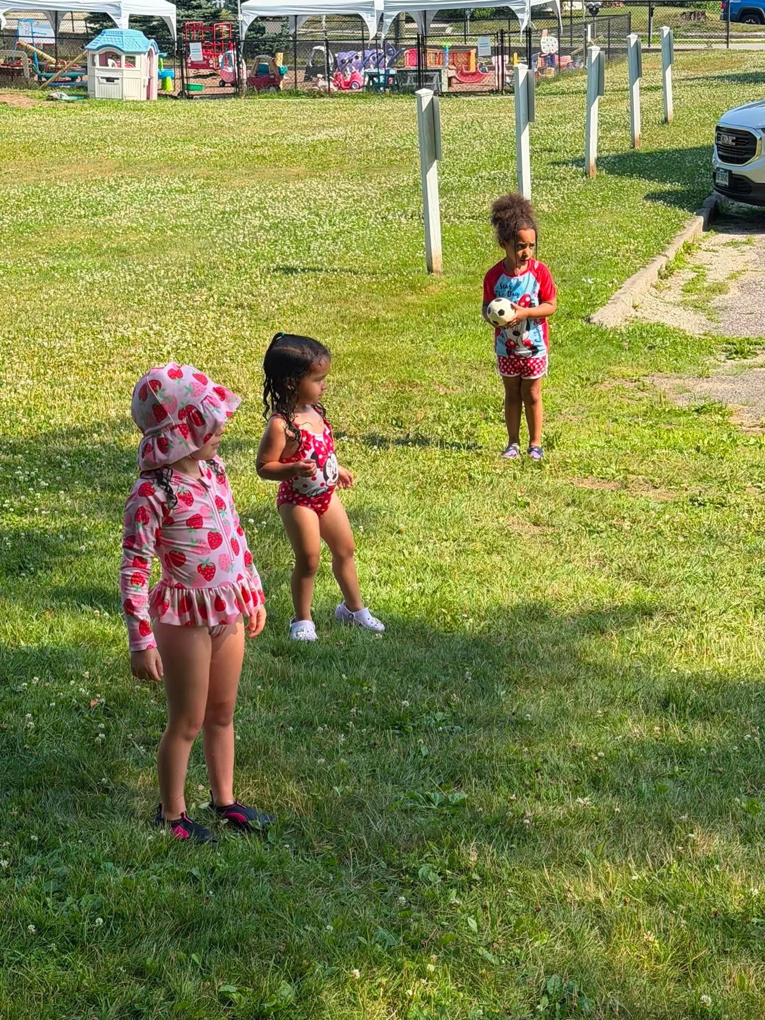 Three children in swimsuits stand on grass near a path at an outdoor park or beach area.