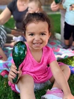 Smiling child in a pink shirt sitting on a blanket, holding a green maraca outdoors.