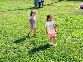 Two children standing on a grassy field, one holding a stuffed toy, with people in the background