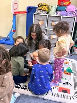 Children gathered around a teacher on a classroom rug, looking at a piano mat and colorful toys.