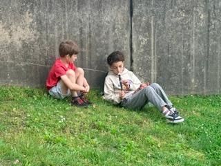 Two children sitting on grass by a gray wall, one in a red shirt and one in a white shirt.