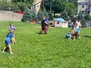 Children playing soccer on a grassy field in a park, with houses and trees in the background.