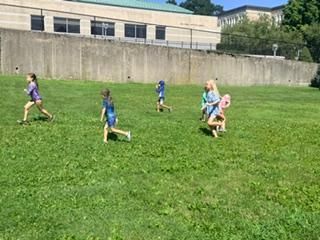 Children running across a grassy field near a concrete wall and buildings