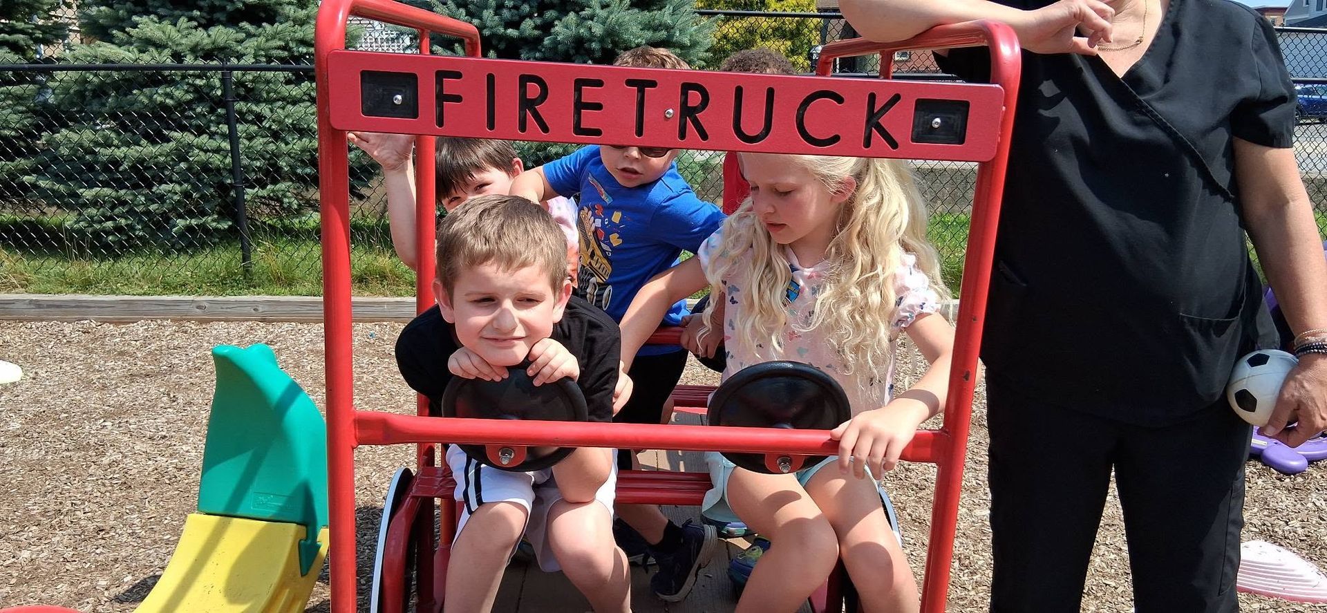 Two children seated in a red firetruck play structure, with an adult standing beside them outdoors.